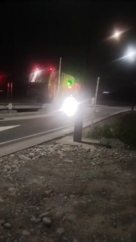 The video captures a nighttime scene at what appears to be a toll booth or checkpoint. A large, bright white light emanates from the center of the frame, likely from a vehicle's headlights. Above this light, a traffic signal displays a green light, indicating that passage is permitted. A barrier arm is partially raised, and behind it, the silhouette of a truck is visible. The truck has red and orange lights on its rear, and its headlights are also contributing to the overall illumination. The road surface is visible, marked with white lines. To the right, a street light illuminates the surroundings. The foreground is composed of rough gravel and stones, with some grassy vegetation visible further back. The sky is dark, with a distinct light source visible in the upper right quadrant of the frame, possibly another street light or a celestial body. The overall atmosphere is dark and industrial, with the focus being on the movement and signaling of vehicles through the checkpoint.