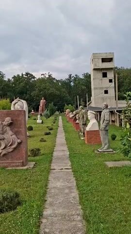 The video pans across an outdoor sculpture park. In the foreground, to the left, is a large, reddish-brown stone relief with figures carved into it. The text on this sculpture is in Cyrillic and reads "НИКОЛАЮ ГРИГОРЬЕВУ". To the right of this relief is a tall, abstract white sculpture of two hands holding a sphere. A concrete path runs down the center of the scene, leading towards a line of busts and statues of historical figures, many of them appearing to be political leaders, arranged on either side of the path. Behind these sculptures, a tall, unfinished concrete building with exposed rebar stands to the right. The background is filled with lush green trees, suggesting a rural or park-like setting. The sky is overcast, indicating a cloudy day. The overall atmosphere is somewhat somber, with the statues of stern-faced individuals and the unfinished building contributing to a sense of melancholy or historical reflection. The video appears to be a casual tour or observation of this outdoor art installation.