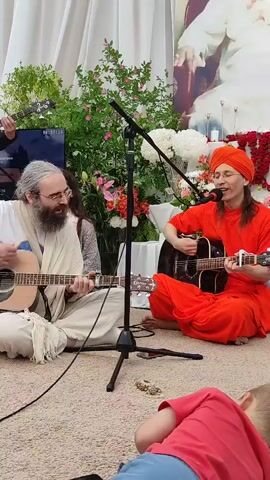 A group of people are gathered in an outdoor setting, possibly a garden or courtyard, for what appears to be a musical performance. Two men are prominently featured in the foreground, both seated on the ground and playing acoustic guitars. The man on the left has a long, grey beard and is wearing a light-colored robe. The man on the right is wearing an orange robe and a matching orange head covering. Both men are singing into microphones positioned in front of them. Behind them, a woman is visible singing along. To the left, a young person plays a bass guitar. In the far background, a large portrait of a religious or spiritual figure is displayed against a white backdrop adorned with flowers and red fabric. A small child wearing a red shirt and blue shorts is lying on the ground in the immediate foreground, facing away from the camera. The ground is covered with a light-colored carpet or mat.