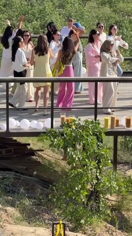 A group of approximately 15 women dance on a wooden deck. The deck is elevated and features white balloons along the edge facing the camera. Clear glasses filled with varying shades of orange and yellow liquid are lined up on the edge as well. The women wear a variety of outfits, including white blouses, a yellow dress, pink pajamas, jeans and a t-shirt, and traditional garments. Most of them wear sunglasses. They appear to be enjoying themselves, moving their arms and bodies to the music. The background features dense green trees and a grassy hillside. A small shrub obscures part of the foreground. A yellow case or box is visible on the ground in the lower left corner. The camera angle is slightly low, looking up at the deck and the women.