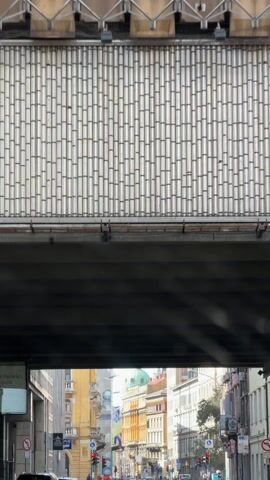The video shows a view from underneath a bridge, looking down a street in a city. The buildings on either side of the street are tall and made of stone or concrete, with different architectural styles. Some buildings have balconies and ornate details. In the distance, a building with a green dome is visible. There are traffic lights at the intersection, and some cars and pedestrians are moving on the street. The sky is blue with a few clouds.