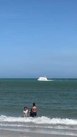 Three children wade in shallow ocean water under a clear blue sky. A small white boat is visible further out in the water. Two of the children are boys, one wearing dark swimming trunks and the other in shorts. The third child is a girl in a white outfit. One of the boys is splashing water. The camera is at the water's edge and captures another person walking into the shot briefly in the foreground. The water has small waves and rocky formations are seen in the distance.