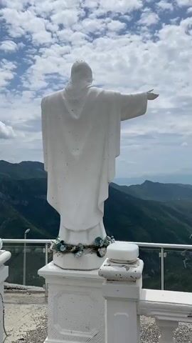 A white statue of Jesus Christ, seen from behind, stands on a viewing platform, its right arm outstretched. The statue has a wreath of blue and white flowers at its base. The camera slowly pans to the right, revealing a breathtaking view of a deep, green mountainous valley under a partly cloudy sky. A winding river and a road are visible at the bottom of the valley, stretching into the distance. A metal and glass railing marks the edge of the platform where the statue is located. The scene is bright, suggesting daytime.