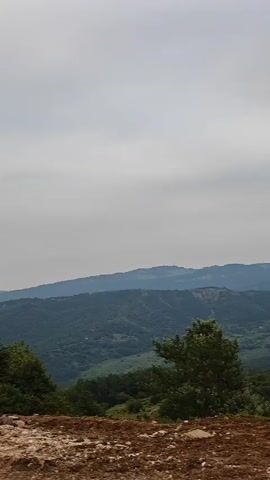 The video shows a tall stone tower on a rocky hill with a background of forested mountains under a cloudy sky. The tower is made of rough-hewn stone blocks and has several small window openings at different levels. There is a doorway at the base of the tower. To the left of the tower, there are trees and a pile of rocks. To the right of the tower, there is a blue object and some greenery. A person is visible near the base of the tower on the left side of the frame. The camera appears to be moving slightly.