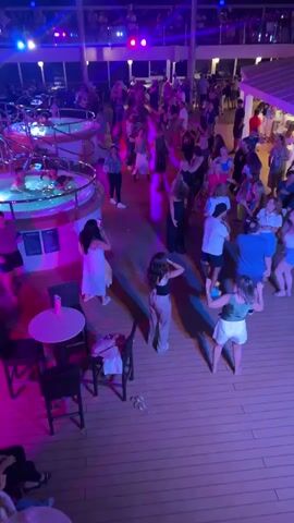 The video shows a crowded outdoor party area at night, illuminated by colorful lights, predominantly pink and blue. A group of people are dancing on a wooden deck, and some are in hot tubs. Several tables and chairs are visible, and a railing separates the deck from a higher level where more people are gathered. The atmosphere appears festive and energetic, with people interacting and enjoying themselves.