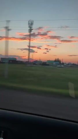 The video shows a car passenger's view of a rural landscape during sunset. The sky is filled with pink and orange clouds, with a clear blue section above. In the distance, there are several houses and a tall cell tower. A green field stretches across the foreground, with a dark road running along the bottom edge of the frame. The camera is moving, suggesting the video was taken from a moving vehicle.