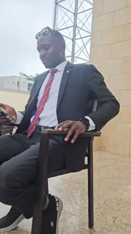 A man wearing a black suit, white shirt, and a striped red tie sits on a black chair with a metal frame. He has dark skin, is wearing glasses on his head, and is looking down at his phone. The background appears to be outdoors with a beige tiled wall to the right and a metal structure in the distance. The lighting suggests it is daytime.