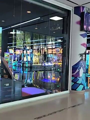 This is a video taken inside a game arcade. The arcade is well-lit with neon lights from various game machines. The entrance to the arcade has a sign that says "GAMESTATION" in large letters. To the left of the entrance, there is a glass window showing more arcade machines inside. To the right of the entrance, there is a pillar decorated with a mural of a cartoon character. The floor is a light-colored tile. The video pans from left to right, showing the exterior of the arcade and the colorful game machines within.
