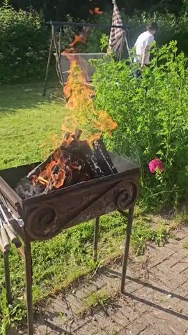 A backyard scene captured in daylight shows a metal grill filled with burning logs. Flames rise from the wood, casting an orange glow. To the left of the grill are several metal skewers with wooden handles. In the background, a person is seated facing away from the camera. The environment includes a lush green lawn, trees, and bushes. There is also a swing set visible in the distance. The overall atmosphere suggests an outdoor gathering or preparation for a barbecue.