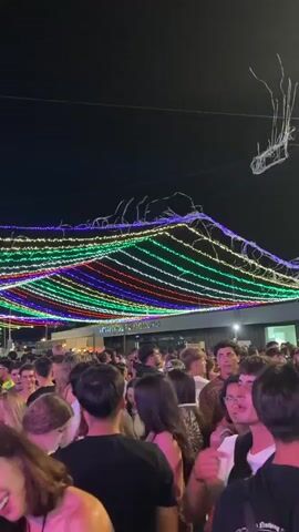 The video shows a crowded outdoor event at night, likely a festival or street fair. Colorful string lights are strung across the scene, creating a festive atmosphere. A large group of people, predominantly young adults, are visible, many of whom appear to be dancing and socializing. The lighting is dim, illuminated by the colorful lights and some ambient sources from buildings that are not fully visible. The overall impression is one of lively celebration and community gathering.