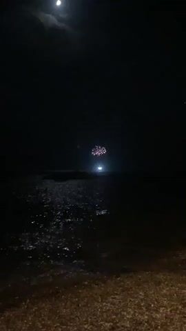 A nighttime video captures a dark sky with a bright moon visible in the upper left corner. Below the moon, across a dark expanse of water reflecting the moonlight, is a bright light, possibly a boat or distant structure. Above this light, fireworks explode in a cluster of colorful bursts. The foreground shows a dark, textured surface, likely a beach or shoreline, with small waves lapping at the edge. The overall scene is one of a nocturnal event with illumination from the moon, man-made lights, and fireworks over the water.