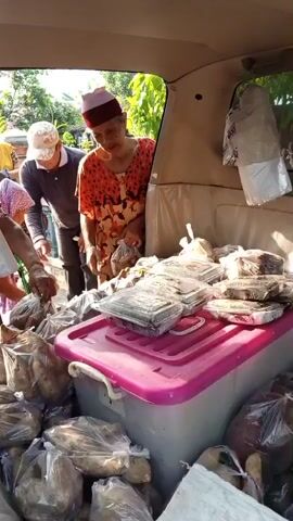 The video shows the back of a vehicle, likely a van or SUV, filled with produce and other items. Several people are gathered around the open rear of the vehicle. In the foreground, there are many large, brown tubers, possibly sweet potatoes or yams, packed in clear plastic bags. They fill the lower portion of the vehicle's cargo area. On top of a large pink and gray plastic bin, there are stacks of clear plastic containers, each containing items wrapped in clear plastic. The containers appear to be food items. One woman, wearing a red and orange patterned dress and a head covering, is holding a bunch of the bagged tubers and looking down at them. Behind her, another person in a gray shirt and a white head covering is also looking down at the produce. Further back, other people are visible, also attending to the items in the vehicle. The environment appears to be outdoors, with sunlight casting shadows and trees visible in the background. The overall scene suggests the loading or unloading of goods, possibly for sale or distribution.