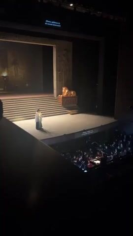 From a high vantage point within a theater, a stage is set to represent an ancient scene, possibly Egyptian-themed, featuring a woman in a long, light-colored robe standing on a broad platform. Behind her, several stairs ascend to a backdrop with artwork and a golden, lion-like sculpture. Below the stage is an orchestra, likely providing live music for the performance. Above the stage, subtitles in various languages are displayed. The room is dimly lit, emphasizing the stage as the primary focal point.