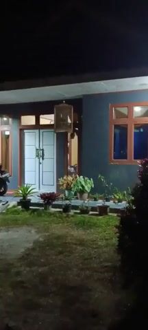 A person walks from right to left across a darkened yard toward a house at night. A motorcycle is parked on the left side of the yard, near the house. The house has a dark gray exterior with a covered porch and a white double door. Several potted plants are arranged in front of the house. In the background, other houses and lights are visible in the distance. The camera is handheld and moves as the person walks.