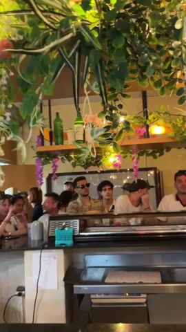 The video shows a busy scene inside what appears to be a restaurant or bar. In the foreground, the camera is positioned behind a counter with a metal surface and a blue plastic organizer containing what looks like cutlery or condiments. There's a receipt dispenser visible on the counter. Behind the counter, several people are gathered. On the left, a woman with dark hair is visible, partially obscured. Further back, a group of men are standing or sitting at a bar. One man in the center is wearing sunglasses and a patterned shirt. Another man on the right is wearing a white shirt. A woman with dark hair, wearing a patterned top and a dark apron, is standing in the middle, facing away from the camera, and appears to be interacting with a tablet or a payment device. Above the bar area, the space is decorated with an abundance of artificial greenery, some pink flowers, and fairy lights, creating a lush and vibrant atmosphere. The background shows more people, a window with a wooden frame, and shelves with bottles and decorative items. The scene is somewhat blurry, particularly in the latter half of the clip as the camera moves, creating a dynamic and lively impression.