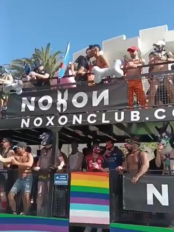 A two-story float filled with people is moving down the street. The float is decorated with the words "NOXONCLUB.COM" and "PROUD OF BEING YOURSELF." Many people on the float are wearing pup masks or other fetish gear.  Some of the people on the top level are shirtless. A person on the street in a pup mask and costume holds a fan. The sky is bright and the weather seems warm.  Other people are walking along the street near the float.