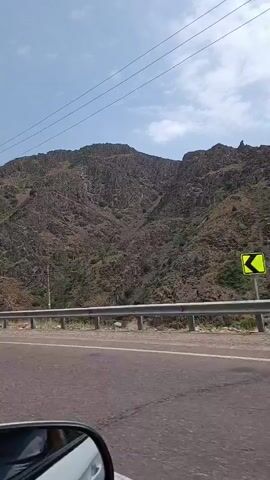 The video shows a view from a car moving along a road. In the foreground, the edge of the car's side mirror and part of the car's body are visible on the left. The road itself is asphalt with white lane markings and a silver metal guardrail separating it from the embankment. The embankment is steep and rocky, covered with sparse vegetation. A tall, slender concrete utility pole with wires attached stands near the road. Further in the background, more utility poles and a taller metal tower are visible. The sky above is a clear blue with a few wispy clouds. The camera appears to be panning slightly to the right as the car moves forward.