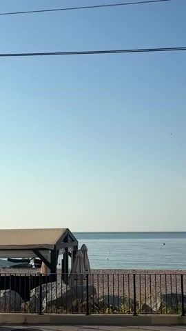 A static, eye-level shot shows a clear blue sky above a calm, blue ocean and a pebble beach. In the foreground, a dark metal fence separates the viewer from the beach. To the left, a covered structure with a light-colored canopy and dark wooden beams stands on the beach. There are two closed umbrellas leaning against the structure. Scattered yellow buoys and two distant figures are visible in the water. A single person is seated on the beach behind the fence. Overhead power lines are visible in the top portion of the frame.