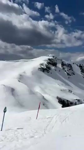The video shows a wide-angle view of a snow-covered mountainside, presumably a ski resort, under a partly cloudy sky. The foreground features a ski slope marked by blue and red poles, indicating trail boundaries or course markers. Ski tracks are visible in the snow, suggesting recent activity. Further up the slope, ski lifts and more trails can be seen, leading towards distant, snow-capped mountain peaks on the horizon. A signpost with an arrow pointing left is also visible, providing directional information for skiers. The overall environment is bright and sunny, typical of a high-altitude ski resort setting.