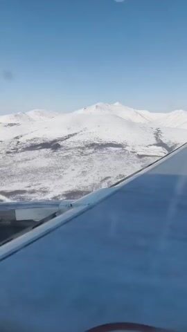 The video shows a view from an airplane window. The wing of the plane is visible, and in the distance, there are snow-covered mountains. The sky is clear and blue. A voice speaks in Russian saying "Our plane is ready to land. We ask you to make sure that all electronic devices are turned off."