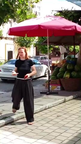 A woman in a black t-shirt and loose black pants walks along a tiled sidewalk. She is holding cash in her right hand and a red object, possibly a phone, in her left. To her right is a fruit stand with watermelons, bananas, tomatoes, and other produce displayed under a red umbrella. Further down the sidewalk, there are more fruit stalls and shops. Cars are visible on the street in the background. The scene appears to be a bustling outdoor market on a sunny day.