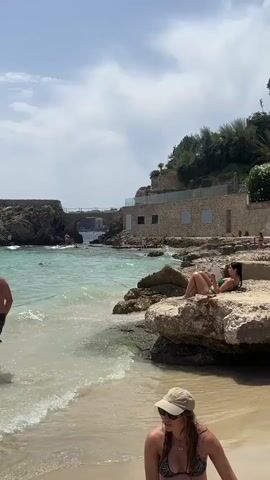 The video shows a beach scene with clear turquoise water and a sandy shore. In the foreground, a person wearing a cap and bikini is partially visible, looking down. In the mid-ground, a woman in a bikini is sitting on rocks by the water, reading a book. Beyond her, there is a stone building with a modern extension and a bridge connecting to a rocky outcrop. Several people are visible in the water and on the shore. The sky is blue with scattered clouds.
