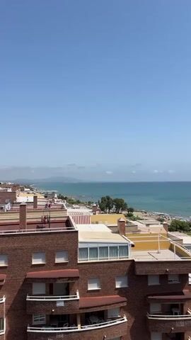 A high-angle shot overlooks a coastal city with buildings in the foreground and the ocean in the background. The sky is clear and blue. The buildings are made of brick and have balconies and rooftops. The ocean is a vibrant blue-green, and a distant coastline is visible. The scene appears to be set on a sunny day.