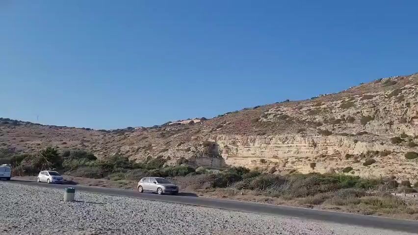 The video shows a daytime view of a road with two cars driving on it. A light-colored sedan is in the foreground on the left side of the road, and a darker sedan is further down the road on the right side. The road is paved and bordered by gravel and sparse vegetation. In the background, a large, rocky hill with layered geological formations dominates the landscape. The sky is clear and blue. The overall scene suggests an arid or semi-arid environment.