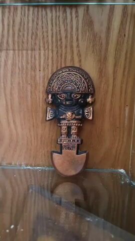 The video shows a close-up view of several decorative objects displayed on wooden shelves, likely within a cabinet. The lighting is somewhat dim. The first object, seen at the beginning of the video, is a small, metallic figurine with a copper-like finish, depicting a stylized human-like figure with a headdress and a base that has the word "PERU" inscribed on it. The camera then pans to the right, focusing on several metallic ornaments shaped like dragons. These dragon ornaments are intricately detailed and have a silver or pewter-like appearance. They are arranged on different shelves, suggesting a collection. The background consists of the textured wooden surface of the shelves and the cabinet. The video is steady, indicating the camera is likely mounted or held still. There are no people or animals visible in the video. The overall impression is of a display of cultural artifacts or decorative items.