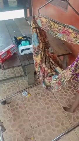 This video shows a child swinging in a hammock. The child, wearing a colorful t-shirt, is a young boy. He is being gently rocked back and forth in a patterned fabric hammock. The hammock is suspended from a metal frame, suggesting it is a portable baby swing or similar apparatus.

The setting appears to be outdoors, possibly a porch or patio, given the tiled floor and the wooden table in the background. The tiled floor has a light brown and beige mosaic pattern. Some objects are visible on the wooden table, including what looks like a red box and some white containers. A few small yellow and white packets are also on the tiled floor.

The activity is simple and peaceful: the child swinging. The motion of the hammock is the primary movement in the video, causing the fabric to sway and creating a slightly blurred effect in some frames. The child's face is mostly obscured, but he appears relaxed, perhaps even sleepy, as he swings.

There is no clear indication of the time of day or weather conditions, though the lighting suggests it is daytime. The environment seems to be in a residential or casual outdoor space. No text is clearly legible within the video. The overall mood is one of quiet observation and a moment of calm for the child. The location context provided is Perdamean, Indonesia.