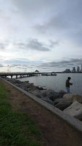 A man fishes from a rocky shoreline with a cityscape and pier in the background.