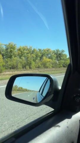 The video is a first-person perspective from inside a car, looking out the side window and into the side mirror. The car is traveling on a paved road. The sky is clear and blue. On the right side of the road, there is a line of trees with green and yellow foliage, indicating it might be autumn. In the side mirror, the road stretches ahead, and another car is visible in the distance, also traveling in the same direction. The interior of the car, specifically the door panel and window frame, is visible in the foreground. The overall scene depicts a car journey on a clear day.