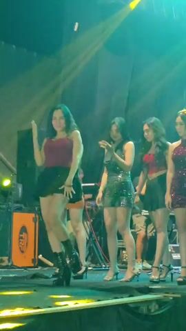 Four young women are on a stage, lit by colorful spotlights. The woman in the front left wears a red camisole and a black mini skirt, paired with black platform boots. She is dancing and smiling. The woman next to her wears a brown halter top and an orange mini skirt, with high heels. She is also smiling. The third woman from the left wears a sparkly silver dress and high heels. She is holding a phone and looking at it. The fourth woman on the right wears a red sequined top and a black skirt with high heels, and she is smiling. There are stage lights, speakers, and other equipment visible on the stage. The background appears to be a dark curtain or wall.