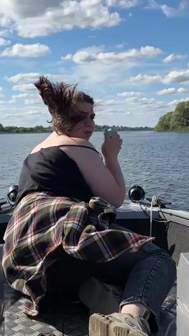 A person is sitting in a boat on a body of water, possibly a lake or river. They are looking over their shoulder towards the camera and smiling. The person is wearing a black tank top, a plaid shirt tied around their waist, and jeans. They are holding a light blue object in their right hand. The boat appears to be moving, as evidenced by the slight blur of the water and the wind blowing the person's hair. The sky is blue with fluffy white clouds, and trees line the distant shore. The overall scene suggests a casual outing on the water on a sunny day.