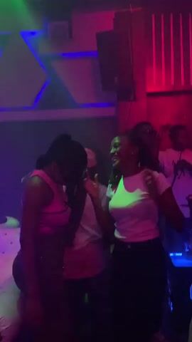 The video captures a dimly lit, vibrant nightclub scene with pulsating colored lights. In the foreground, two women are the primary focus. The woman on the left is wearing a pink tank top and jeans, with her hair pulled back in a high ponytail. She appears to be interacting with the woman on the right, who is wearing a white t-shirt and dark pants. The woman on the right has braided hair and is looking down, seemingly engaged in an activity. Between them, a person wearing a white t-shirt and a pink baseball cap is partially visible. In the background, other individuals can be seen, some dancing. The environment features nightclub decor, including geometric wall panels illuminated with blue and green lights, and a red light fixture on the right. The overall atmosphere suggests a lively social gathering.