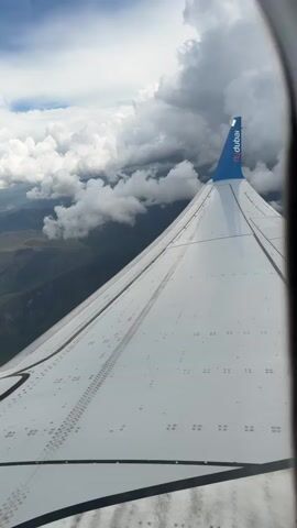 From an airplane window, a view of the wing of a flydubai aircraft fills the lower half of the frame. The wing is white and features rows of rivets. The tip of the wing displays the flydubai logo in blue. Above the wing, the sky is visible, featuring patches of blue interspersed with puffy white and gray clouds. Below the clouds, a landscape of green hills and mountains stretches towards the horizon. A city is visible in the distance, partially obscured by the clouds. The scene is bright and daytime. Sounds of children talking can be heard in the background.