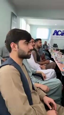 In an indoor setting, likely a meeting or conference room in Shurandam, Afghanistan, several men are seated and appear to be attending a presentation or lecture. The room has light-colored walls and windows, indicating daytime.

In the foreground, a young man with dark hair and a beard is seated, facing forward with a serious and attentive expression. He is wearing a light brown long-sleeved shirt under a dark blue vest. Behind him, several other men are also seated, some wearing traditional Afghan attire such as the Salwar Kameez. They too are looking forward, suggesting they are focused on a speaker or presentation not visible in the frame. An older man is discernible further back in the room.

A long table is visible in front of the attendees, holding glasses and bottles. In the background, a white banner or screen displays a logo with the letters "AC" clearly visible, along with some text in an apparent script (likely Dari or Pashto) and partial English text that seems to include "Chamber of Co...". This suggests an event related to a Chamber of Commerce or similar business organization. The overall atmosphere appears professional and focused.
