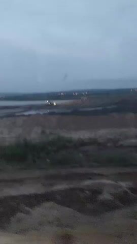 The video begins with a wide shot of a desolate landscape under a cloudy sky. In the distance, there are lights visible, possibly from buildings or a town. A body of water can be seen on the left side of the frame, and what appears to be a construction or excavation site is spread out in the midground. Several pieces of heavy machinery, possibly excavators or loaders, are active at the site, their lights illuminating the dark ground. The camera then shifts to a close-up of a side-view mirror of a vehicle, reflecting the dim, yellowish light from the headlights of another vehicle. The next shot is from inside the cabin of a heavy vehicle, likely a loader or a truck, showing a dirt road ahead. The headlights of the vehicle are on, illuminating the road and the surrounding sparse vegetation. The dashboard of the vehicle is visible in the foreground, with various illuminated gauges and indicators. One gauge shows a speed of approximately 30 kilometers per hour. The sky above is a dark, dusky blue, with hints of purple and orange, suggesting either dawn or dusk. The video ends with a focus on the illuminated dashboard.