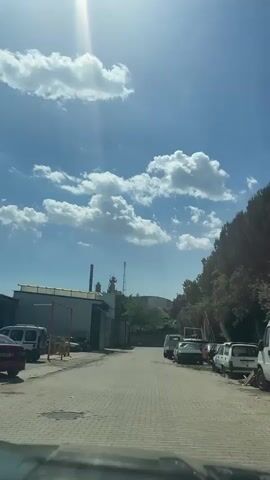 The video shows a street view under a bright blue sky filled with puffy white clouds. The sun shines brightly, creating a lens flare effect. The street is paved with interlocking bricks, and on the left side of the frame, there are several industrial-looking buildings with closed roller doors. Some cars and trucks are parked along the right side of the street, next to a tree-covered slope. A telecommunications tower is visible further down the street in the distance. The camera is positioned inside a vehicle.