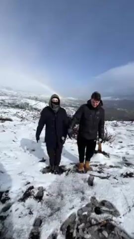 Two people stand on a snowy hilltop under a partly cloudy sky with a rainbow visible in the distance. The person on the left wears a dark coat, brown pants, a dark scarf covering their lower face, and dark shoes. They hold a beverage can in their left hand. The person on the right wears a black puffer jacket, black pants, and tan work boots. They initially hold their right hand to their head in a salute-like gesture, then lower it. The ground is covered in snow with patches of bare earth and small branches visible. Footprints are visible in the snow in the foreground.