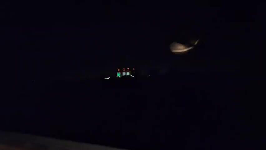 The video captures a night scene with a dark sky. In the center of the frame, a building is illuminated with colored lights. The lights appear to be arranged in vertical columns, with green lights on the left, followed by white lights in the middle, and red lights on the right. The building itself seems to be a large structure, possibly industrial, with multiple levels or sections. In the foreground, the edge of a vehicle's interior is visible, suggesting the footage is from inside a car. There are a few other distant lights scattered across the landscape, indicating a sparsely populated area or the outskirts of a town. A small, bright light, possibly another distant light source or a reflection, is visible to the right of the building. The overall impression is one of a nighttime drive passing by a lit structure.