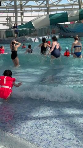The scene is an indoor water park where multiple people, including adults and children, are enjoying themselves in a wave pool. The pool is filled with artificially generated waves, and the bottom has a patterned tile design with hues of blue and white. Several individuals are wearing life vests, mainly children. A group of people can be seen wading through the water, while others are standing and enjoying the waves. In the background, there's a large, green and white water slide and structural supports of the water park's ceiling. The scene appears to be well-lit with natural light streaming in through the roof. There is signage with the Russian words "АКВАПАРК На".
