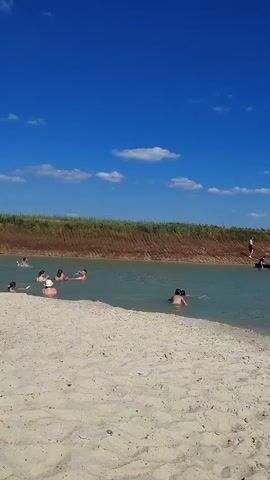 This media file depicts a sunny day at a sandy beach with a clear blue sky and scattered white clouds. The scene is a natural body of water, likely a lake or pond, with calm, light blue-green water. The shoreline is sandy, with some beach toys, including a pink bucket and a blue watering can, visible on the sand.

Several people are enjoying the water. In the foreground, a young child in a colorful swimsuit and pink headscarf is running on the beach. Further back in the water, other individuals are swimming or floating. Some are partially submerged, with only their heads visible, while others are more fully in the water. On the right side of the frame, a person is partially visible on an inflatable tube in the water, and another person is standing on the embankment behind them. The embankment is covered in dry grass and reddish-brown earth.

The time of day appears to be midday or early afternoon, given the bright sunlight casting shadows on the sand. The weather is clearly warm and pleasant, ideal for swimming and outdoor activities. The setting suggests a recreational area, possibly a less developed beach or a natural swimming spot. The overall mood conveyed is one of leisure and enjoyment on a summer day. There is no visible text in the media file.