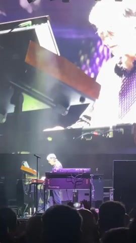 A man with graying hair is shown playing a keyboard on a stage. He is wearing a patterned shirt and is looking down at the keyboard. In the background, a large screen displays a close-up of the man's face, also looking down. Another smaller screen is visible to the left of the stage, displaying a portion of the man's hands on a keyboard. The stage is lit with purple and white lights, and a crowd of people is visible in the foreground, holding up their phones. The overall impression is of a live musical performance.