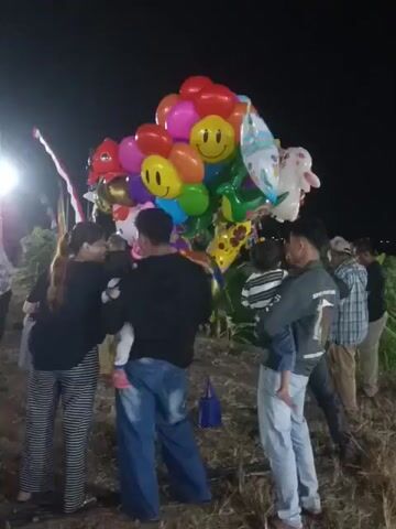 A nighttime outdoor scene with a cluster of brightly colored balloons, including some shaped like smiley faces and cartoon characters, held by an unseen person. Several people are gathered around the balloons, some holding children. One person on the left is wearing a black top and striped pants, holding a baby. To their right, a person in a black hoodie and blue jeans is also holding a baby. Further to the right, another person in a denim jacket and light blue jeans is holding a child. In the background, there is a crowd of people, illuminated by several bright lights, suggesting a lively event or market. The ground appears to be dirt or dry grass, and some foliage is visible.