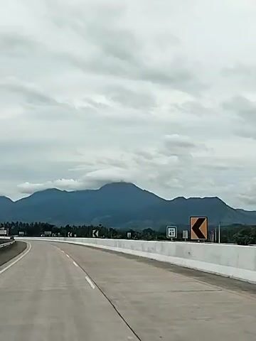 The video shows a drive on a highway. The road is paved and has a white line marking the center. There is a white barrier on the right side of the road, with yellow and black curve warning signs. In the distance, there are blue mountains under a cloudy, grey sky. The scenery appears to be rural, with trees visible in the background. The camera seems to be mounted inside a moving vehicle, capturing the perspective of the driver.