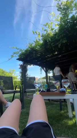 This video shows a group of people outdoors under a pergola covered in green foliage. The sky is blue with some wispy clouds. In the foreground, a person's legs are visible, wearing black shorts with white trim and white sneakers. A white table is in front of them, with several glasses of drinks and other items on it. In the background, under the pergola, two men are standing and interacting. One man is wearing a white shirt and the other is wearing a grey shirt and shorts. The ground is covered in grass. The scene appears to be a casual outdoor gathering or party.