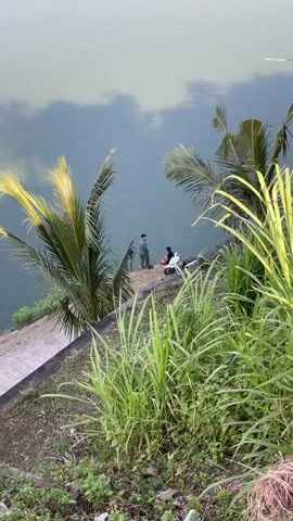 From an elevated perspective, two individuals are visible at the edge of a greenish-blue body of water, possibly a lake. One person is in a dark top and light pants, seemingly leaning towards the water, while the second is in a lighter top and dark pants, seated slightly behind. A white scooter is parked next to them on a small, flat area. To the left, a concrete slope leads down towards the water, with two palm trees growing alongside. The foreground and right side of the frame are dominated by tall, vibrant green grass. Cloud reflections are visible on the water's surface.