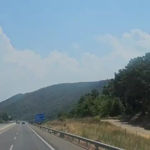 The video shows a highway scene with a bright blue sky and fluffy white clouds. On the highway, there are several vehicles including a blue truck with a white trailer, a white truck, and some cars. The truck with the white trailer is in the foreground and appears to be moving. On either side of the highway, there are green trees and a hilly landscape. The road is paved and has a metal barrier along the side. There are also some road signs visible. The lighting suggests it is daytime.