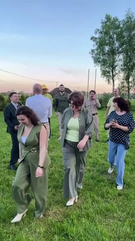 A group of people are standing and walking in a grassy field. Some people are dressed in suits or dress shirts and pants, while others are dressed more casually. One man is wearing a yellow hat. A woman in a polka dot shirt is looking at her phone. One man appears to be holding a microphone. There are houses and trees in the background. The sky is blue with a hint of pink from the sunset.